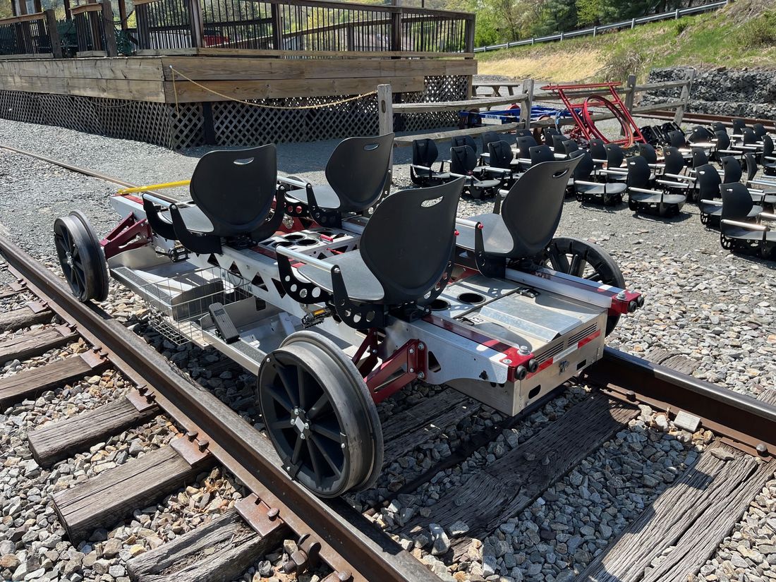 Rear three-quarter view of a Rail Glider showing the industrial aluminum frame, red structural accents, wire baskets, and four flanged rail wheels.