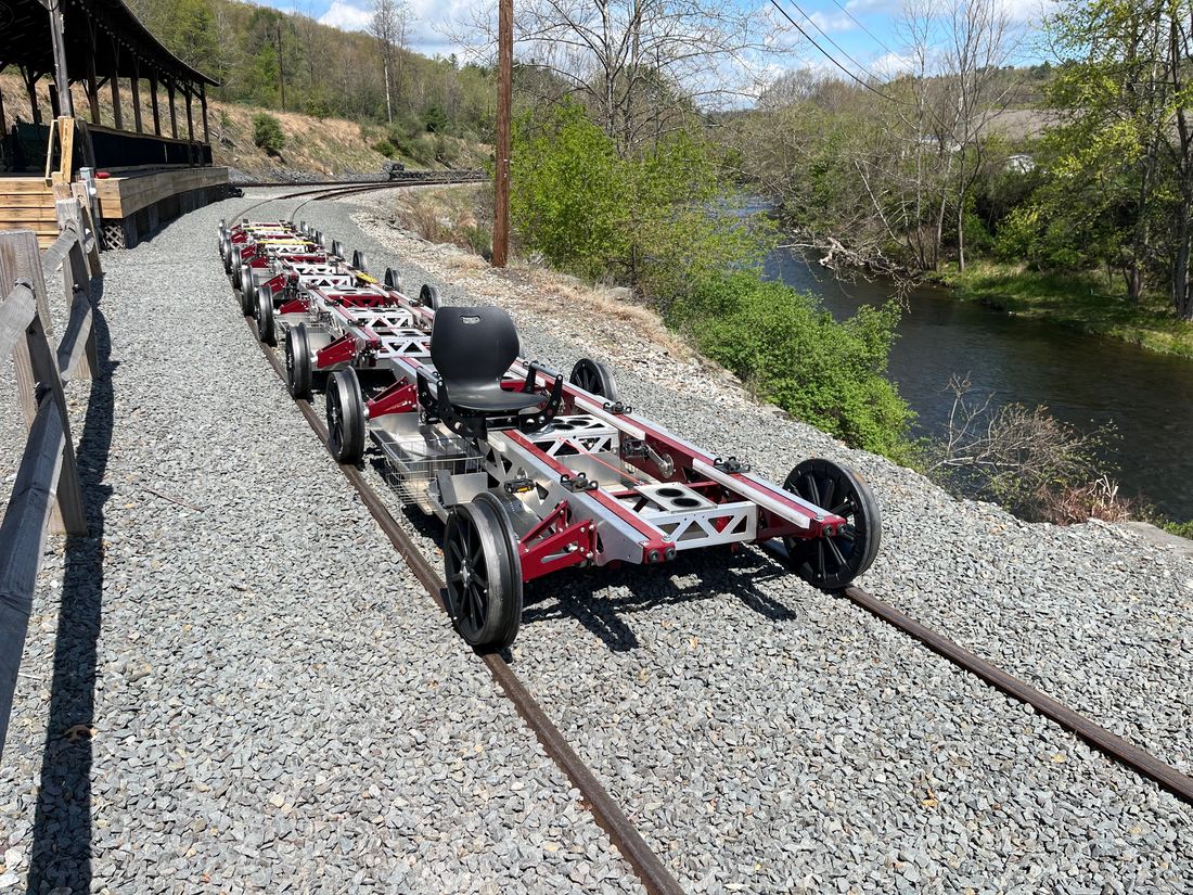 A line of Rail Gliders coupled on the rails beside a river and boarding platform, showing how bikes can run as a train.