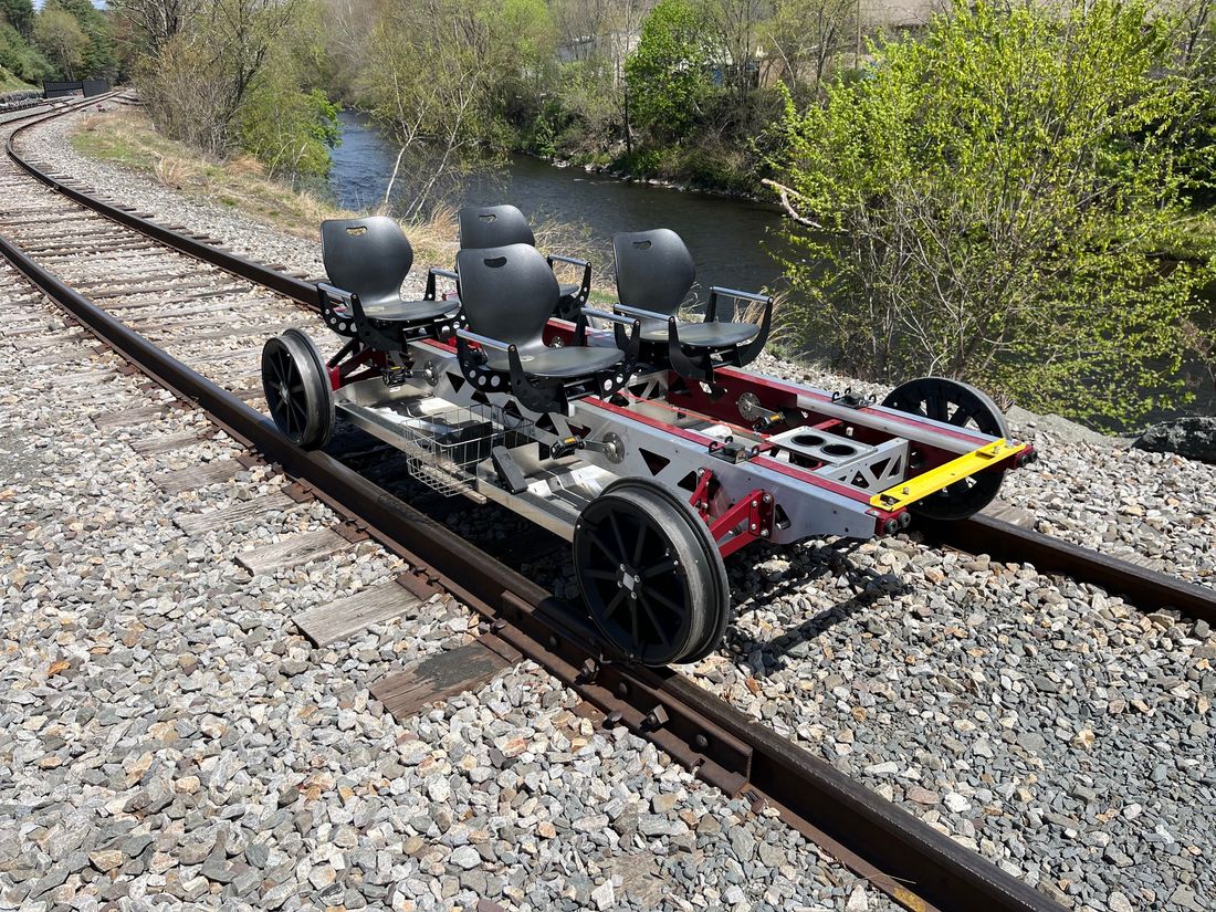 Side view of a Rail Glider on tracks beside a river, showing pedal cranks at each seat and the truss-style side rails.